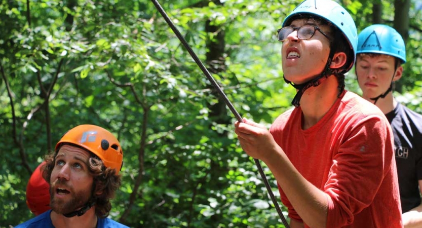 Three people wearing helmets look up at an apparent rock climber while standing in a wooded area. One of them appears to be belaying the climber. 